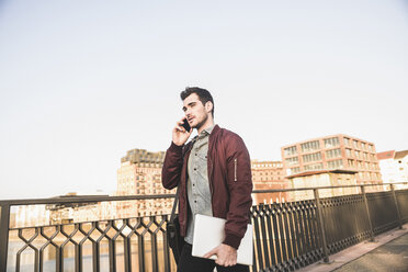 Junger Mann mit Tablet im Gespräch mit Handy auf einer Brücke in der Stadt - UUF16801