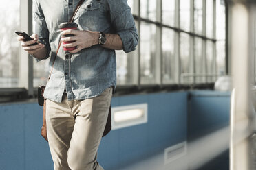 Nahaufnahme eines Mannes mit Handy und Kaffee zum Mitnehmen beim Gehen am Bahnhof - UUF16805