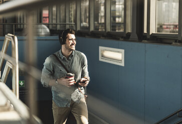 Lächelnder junger Mann mit Kopfhörern, Mobiltelefon und Kaffee zum Mitnehmen beim Spaziergang am Bahnhof - UUF16806