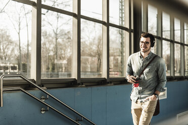 Smiling young man with headphones and takeaway coffee walking at the station - UUF16808