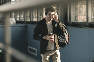 Lächelnder junger Mann mit Kopfhörern, Mobiltelefon und Kaffee zum Mitnehmen beim Spaziergang am Bahnhof - UUF16809