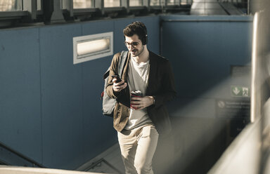 Lächelnder junger Mann mit Kopfhörern, Mobiltelefon und Kaffee zum Mitnehmen beim Spaziergang am Bahnhof - UUF16810