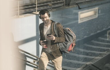 Lächelnder junger Mann mit Kopfhörern, Mobiltelefon und Kaffee zum Mitnehmen beim Spaziergang am Bahnhof - UUF16811