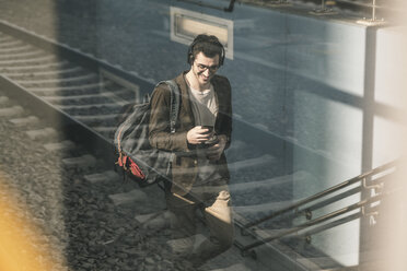 Lächelnder junger Mann mit Kopfhörern, Mobiltelefon und Kaffee zum Mitnehmen beim Spaziergang am Bahnhof - UUF16812