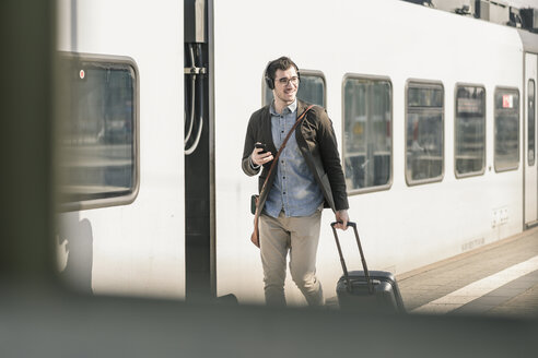 Lächelnder junger Mann mit Kopfhörern, Mobiltelefon und Koffer auf dem Bahnsteig - UUF16817