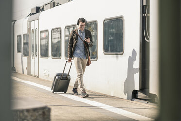 Lächelnder junger Mann mit Kopfhörern, Mobiltelefon und Koffer auf dem Bahnsteig - UUF16818