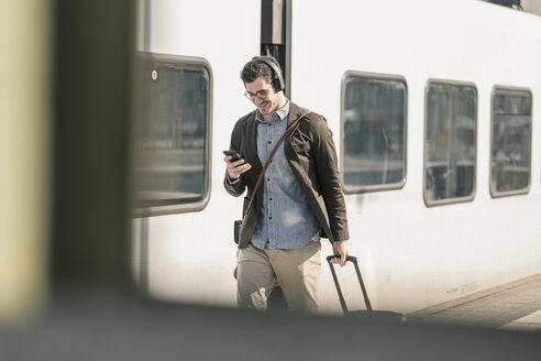 Lächelnder junger Mann mit Kopfhörern, Mobiltelefon und Koffer auf dem Bahnsteig - UUF16819