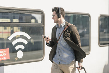 Lächelnder junger Mann mit Kopfhörern und Handy geht entlang des Zuges mit Wifi-Symbol - UUF16820