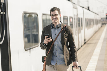 Lächelnder junger Mann mit Mobiltelefon am Bahnsteig - UUF16821