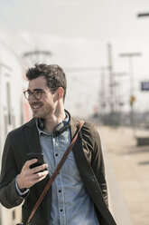 Lächelnder junger Mann mit Mobiltelefon am Bahnsteig - UUF16823