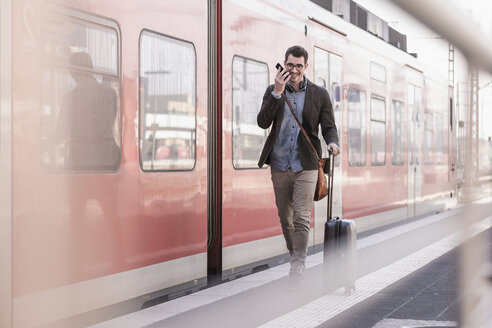 Happy young man with cell phone walking on station platform along commuter train - UUF16831