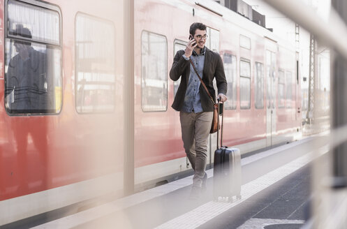 Smiling young man on cell phone walking on station platform along commuter train - UUF16832