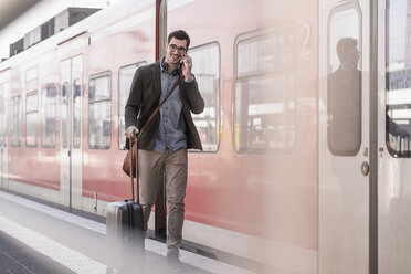 Lächelnder junger Mann mit Handy auf dem Bahnsteig entlang eines Nahverkehrszugs - UUF16833