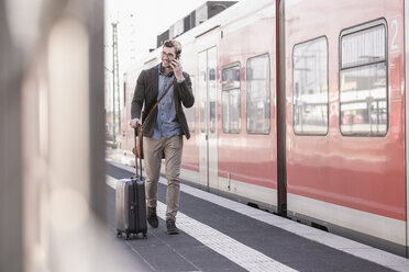 Lächelnder junger Mann mit Handy auf dem Bahnsteig entlang eines Nahverkehrszugs - UUF16835