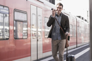 Fröhlicher junger Mann mit Handy auf dem Bahnsteig entlang eines Nahverkehrszugs - UUF16836