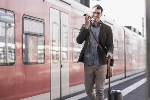 Happy young man with cell phone walking on station platform along commuter train - UUF16836