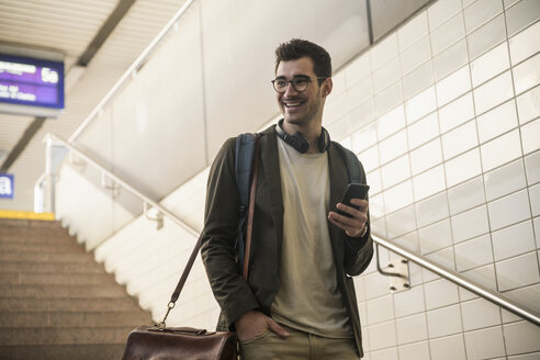 Lächelnder junger Mann mit Mobiltelefon, der die Treppe am Bahnhof hinuntergeht - UUF16838
