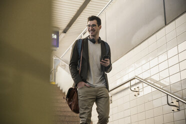 Lächelnder junger Mann mit Mobiltelefon, der die Treppe am Bahnhof hinuntergeht - UUF16839
