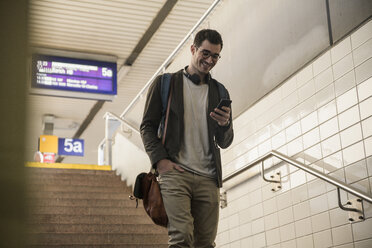 Lächelnder junger Mann mit Mobiltelefon, der die Treppe am Bahnhof hinuntergeht - UUF16841