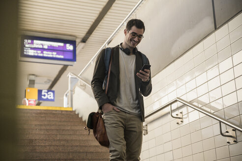 Lächelnder junger Mann mit Mobiltelefon, der die Treppe am Bahnhof hinuntergeht - UUF16841