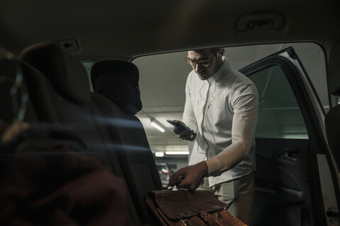Young man with cell phone and earphones taking bag out of car stock photo