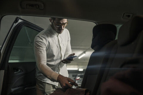 Junger Mann mit Handy und Kopfhörern nimmt Tasche aus dem Auto, lizenzfreies Stockfoto