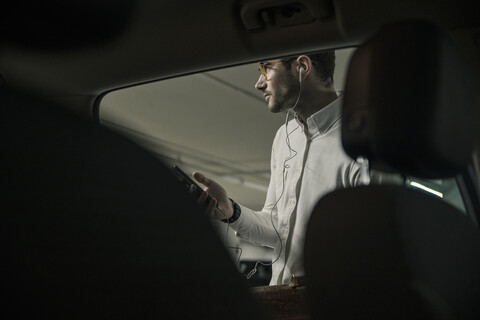 Junger Mann mit Handy, Kopfhörer und Tasche an einem Auto, lizenzfreies Stockfoto