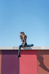 Young woman sitting on a wall listening music with headphones on her smartphone - AFVF02607