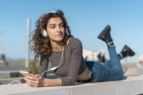Junge Frau liegt an einer Wand und hört mit Kopfhörern auf ihrem Smartphone Musik, lizenzfreies Stockfoto