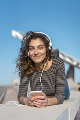 Junge Frau liegt an einer Wand und hört mit Kopfhörern auf ihrem Smartphone Musik - AFVF02610