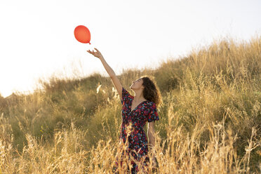 Glückliche junge Frau auf einer Sommerwiese, die einen roten Luftballon loslässt - AFVF02641