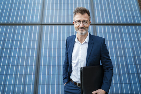 Portrait of smiling businessman holding folder standing in front of solar panels - DIGF06346