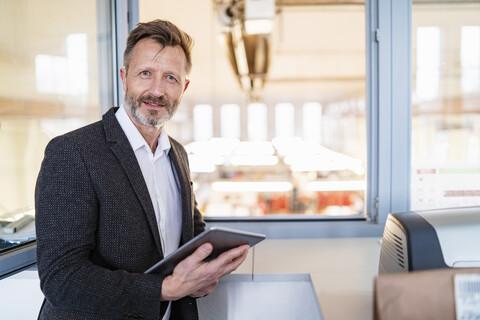 Porträt eines Geschäftsmannes, der ein Tablet am Fenster mit Fabrikhintergrund benutzt, lizenzfreies Stockfoto