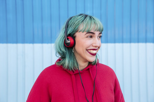 Porträt einer lachenden jungen Frau mit blau gefärbtem Haar, die mit Kopfhörern Musik hört - LOTF00062