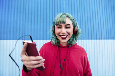 Porträt einer lachenden jungen Frau mit blau gefärbtem Haar und Kopfhörern, die ein Selfie mit einem Smartphone macht - LOTF00065