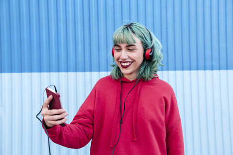 Portrait of laughing young woman with blue dyed hair with headphones taking selfie with mobile phone stock photo