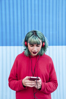 Portrait of young woman with blue dyed hair listening music with headphones looking at smartphone - LOTF00067