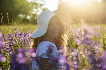 Porträt einer lächelnden Frau mit Sommerhut auf einer Blumenwiese in der Abenddämmerung - SARF04173