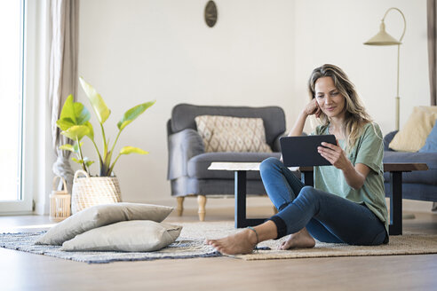 Smiling woman sitting on the floor at home using tablet - SBOF01914