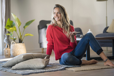 Smiling woman sitting on the floor at home holding cell phone stock photo