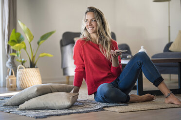 Smiling woman sitting on the floor at home holding cell phone - SBOF01916