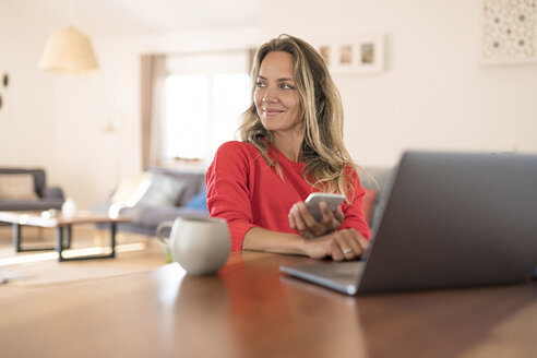 Lächelnde Frau mit Laptop und Mobiltelefon auf dem Esstisch zu Hause - SBOF01935