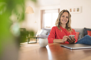 Portrait of smiling woman with laptop and cell phone on dining table at home - SBOF01937