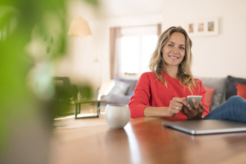 Porträt einer lächelnden Frau mit Laptop und Mobiltelefon am Esstisch zu Hause - SBOF01937