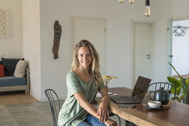 Portrait of smiling woman with laptop sitting at dining table at home - SBOF01962