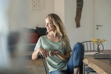 Smiling woman with cell phone and laptop on dining table at home - SBOF01964