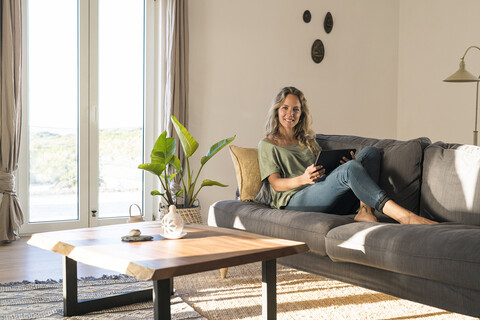Porträt einer lächelnden Frau, die auf der Couch zu Hause mit einem Tablet sitzt, lizenzfreies Stockfoto