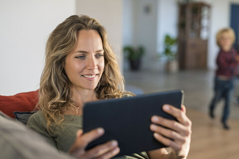 Lächelnde Frau, die zu Hause auf der Couch sitzt und ein Tablet mit ihrem Sohn im Hintergrund benutzt, lizenzfreies Stockfoto