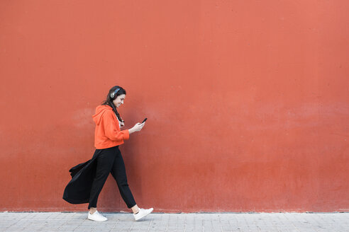 Young dancer walking and using the phone and headphones in front of a red wall - JRFF02886