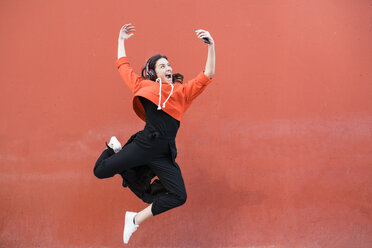 Young contemporary dancer jumping and using smartphone in front of a red wall - JRFF02887